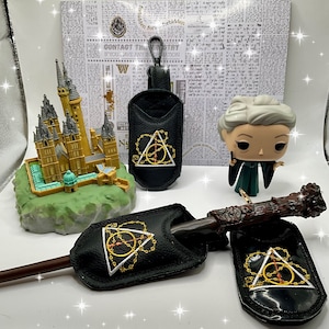 May include: A set of three black leather pouches with gold embroidery of the Deathly Hallows symbol. The pouches are for a wand, a keychain, and a phone. The pouches are displayed on a white surface with a miniature Hogwarts castle model and a figurine of a witch.