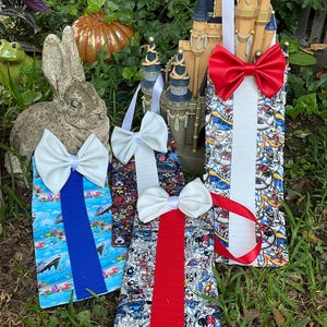 May include: Four hanging fabric towel holders featuring colorful cartoon character prints, each topped with a large solid-colored bow. The towels are displayed vertically, with one featuring a blue strap and white bow, two with white straps and white bows, and one with a white strap and red bow.