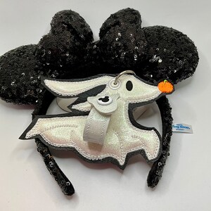 Może przedstawiać: Czarna opaska na głowę z uszami Minnie Mouse z cekinami, do kt&oacute;rej przymocowany jest biały, błyszczący urok Zero, psa-ducha. Urok ma małą dynię i głowę Myszki Miki. Etykieta głosi "Tokyo Disney Resort".