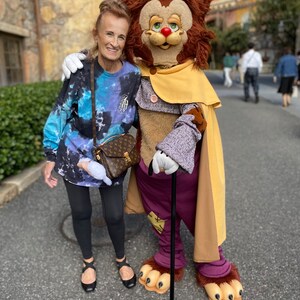 May include: A woman in a blue and white tie-dye long-sleeve shirt and black leggings poses with a furry, brown, and yellow character wearing a purple coat, a yellow cape, and a blue top hat. The character is holding a black walking stick.