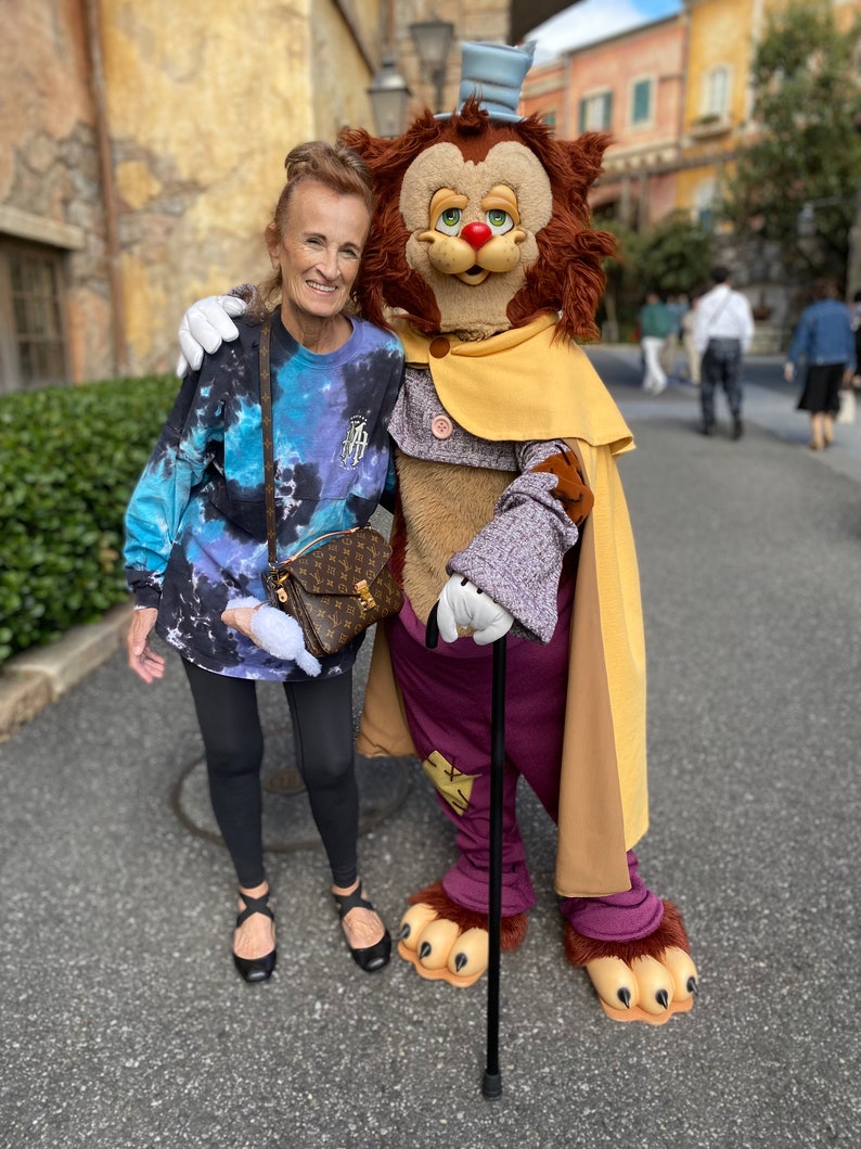 May include: A woman in a blue and white tie-dye long-sleeve shirt and black leggings poses with a furry, brown, and yellow character wearing a purple coat, a yellow cape, and a blue top hat. The character is holding a black walking stick.