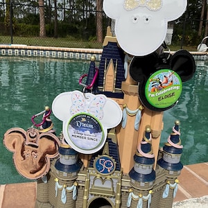 May include: A miniature replica of Cinderella's Castle with a 50th anniversary logo.  There are four Mickey Mouse head-shaped charms hanging from the castle.  One charm is white with a pink bow and sprinkles, another is white with a yellow bow, another is black, and the last is copper.  The charms have various Disney Vacation Club pins attached to them.