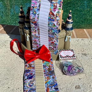May include: A red bow tie with a white and gold striped bow tie with a Disney Parks print. The bow tie is on a white background with a blue pool in the background. The bow tie is part of a set of Disney Parks accessories.