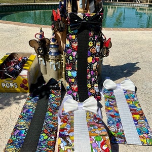 May include: Three colorful fabric straps with black, white, and glitter trim. Each strap features a different Disney character print. The straps are displayed on a table with a box of candy and a toy castle in the background.