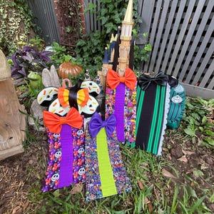 May include: A collection of four vertical fabric hanging organizers featuring various patterns including cartoon characters, stripes, and skulls. Each organizer is topped with a large, solid-colored bow in orange, purple, or black, and includes a hanging loop.