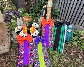 May include: A collection of four vertical fabric hanging organizers featuring various patterns including cartoon characters, stripes, and skulls. Each organizer is topped with a large, solid-colored bow in orange, purple, or black, and includes a hanging loop.