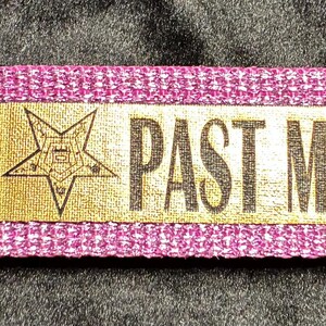 May include: A magenta key fob with a silver-colored metal clasp and ring. A rectangular gold-colored patch is centered on the fob, displaying a five-pointed star and the words "PAST MATRON" in dark brown lettering.