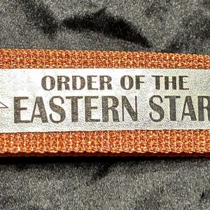 May include: A copper-colored fabric keychain with a silver metal clasp and ring. The keychain features a rectangular silver label with the text "ORDER OF THE EASTERN STAR" and a star symbol.
