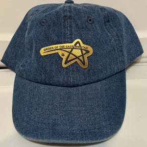 May include: A blue denim baseball cap featuring the text "ORDER OF THE EASTERN STAR" in gold lettering above a gold star emblem. The cap has a curved brim and a button on top.