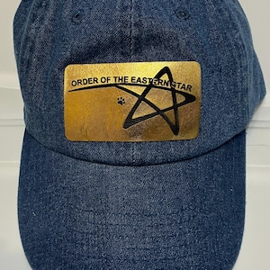 May include: A blue denim baseball cap with a curved brim. The cap features a gold-colored patch with the text "ORDER OF THE EASTERN STAR" and a black star emblem. The cap is a classic style.