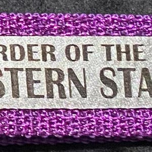 May include: A purple fabric keychain wristlet with a silver metal clasp and key ring. The keychain features a silver rectangular tag with the words "ORDER OF THE EASTERN STAR" and a five-pointed star symbol.