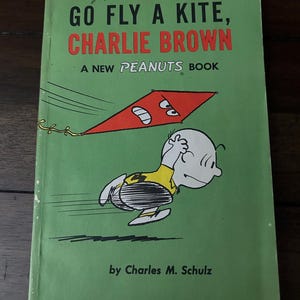 May include: A green cover of a Peanuts book titled "Go Fly a Kite, Charlie Brown" with a red kite and Charlie Brown running with the kite string in his hand. The book is a new Peanuts book by Charles M. Schulz and published by Holt, Rinehart and Winston, Inc.