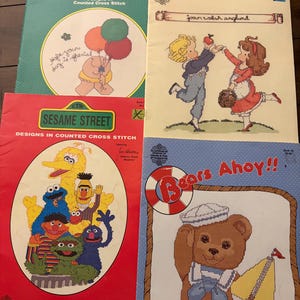 May include: Four vintage counted cross stitch pattern booklets featuring Ziggy, Joan Walsh Anglund characters, Sesame Street Muppets, and a teddy bear titled Bears Ahoy. Each softcover booklet displays colorful illustrations and project designs.