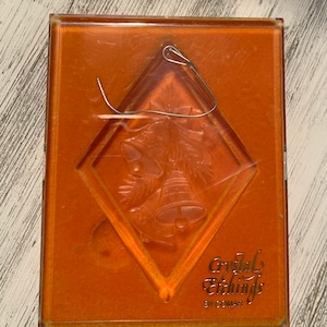 May include: A clear plastic diamond-shaped ornament with a raised design of bells and holly. The ornament is in a brown plastic frame with the text "Crystal Etchings by Comar" printed on the bottom.