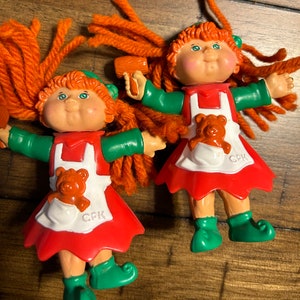May include: Two Cabbage Patch Kids dolls with red yarn hair, wearing green and red dresses with white aprons and a teddy bear pocket. The dolls are holding a wooden mallet and a hair dryer.