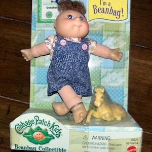 May include: Cabbage Patch Kids Beanbag Collectible toy, a brown-haired doll wearing a blue denim overall dress and a white shirt with pink buttons. The doll is holding a yellow horse beanbag toy. The packaging features a birth certificate and the text "I'm a Beanbag!"