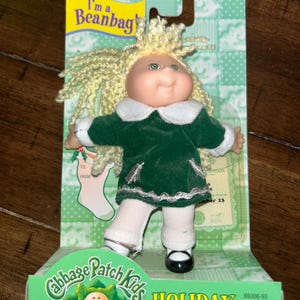 May include: A Cabbage Patch Kids Holiday Beanbag Collectible doll in a green dress with white trim and black shoes. The doll has blonde hair and green eyes. The packaging features the Cabbage Patch Kids logo and the text "Holiday Beanbag Collectible".