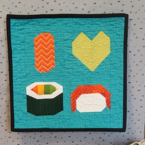 May include: A teal blue quilted fabric square with a black border. The fabric square has four appliqued shapes: a yellow heart, a red and white sushi roll, a green sushi roll with rainbow colored filling, and an orange and white sushi roll. The fabric square is sitting on a gray and white patterned surface.