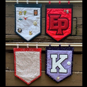 May include: Four rectangular fabric pin banners with pointed bottoms, hanging from wooden dowels. One is white with a dark blue border, one is red with a large red varsity letter Ep, one is white with red stars and a red border, and one is purple with a large white letter K.