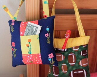 Op de afbeelding: Twee kleine stoffen tassen met banden die aan een houten hoofdeinde hangen. De tas aan de linkerkant heeft een patchwork ontwerp met bloemen en een zakje waarin een dollarbiljet zit. De tas aan de rechterkant heeft een groene en gele stof met een patroon van voetballen.