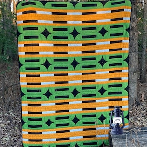 Peut inclure: Une courtepointe colorée avec un motif géométrique de rayures et de formes vertes, noires, orange et blanches. La courtepointe est suspendue à l'extérieur, avec une lanterne bleue vintage sur une surface en bois au premier plan. Décoration intérieure.