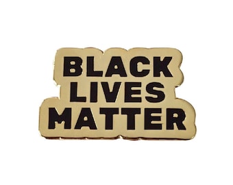 May include: A gold-colored pin with the words "BLACK LIVES MATTER" in bold, black capital letters. The pin has a rounded rectangular shape with a slightly raised border.