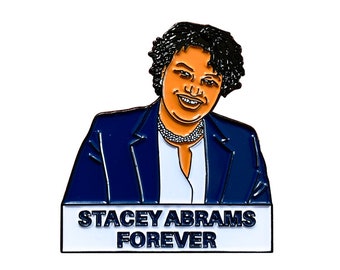 May include: A blue enamel pin featuring a cartoon portrait of Stacey Abrams with the text "Stacey Abrams Forever" on a white banner.