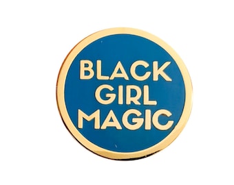 May include: A round, gold-colored pin with a blue center. The text "BLACK GIRL MAGIC" is displayed in white, bold letters. The pin has a shiny, metallic finish and is set against a white background.