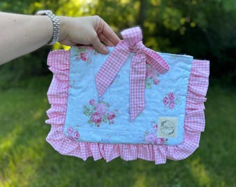 May include: A blue and white floral fabric pouch with a pink and white gingham bow and ruffle trim. The pouch has a small white label with the text "Little Love" on it.