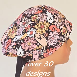 May include: A scrub cap featuring a black background with a repeating pattern of white and peach-colored ghosts interspersed with pink, yellow, and white daisies. The cap includes a black ribbon tie at the back and the text over 30 designs.