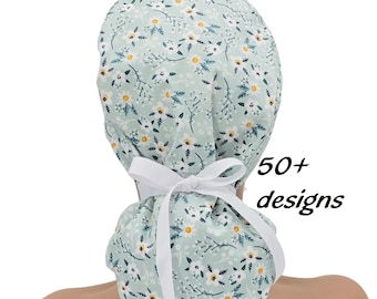 May include: A scrub cap in a light mint green fabric featuring a repeating pattern of white daisies with yellow centers and dark green foliage. The cap includes a white ribbon tied in a bow at the back. The text 50+ designs is visible.
