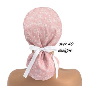 Op de afbeelding: Een oudroze operatiemuts met een wit lijntekening-bloemenpatroon en een wit lint aan de achterkant. De stof heeft een subtiele gespikkelde textuur. De tekst "over 40 designs" staat in het zwart rechts naast de operatiemuts.