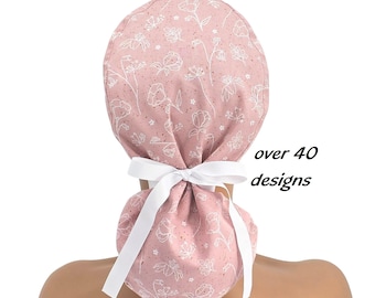 May include: A dusty pink scrub cap featuring a white line-art floral pattern and a white ribbon tie at the back. The fabric has a subtle speckled texture throughout. The text "over 40 designs" is printed in black to the right of the scrub cap.