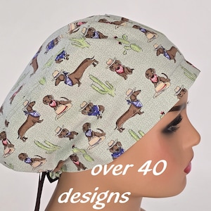 Puede incluir: Un gorro quirúrgico verde claro con un patrón repetido de perros salchicha de dibujos animados con sombreros de vaquero y pañuelos, con ilustraciones de cactus. El texto "over 40 designs" es visible.