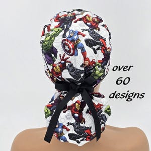 May include: A white scrub cap featuring a colorful print of various Marvel comic book characters, including Captain America, Iron Man, and the Hulk. The cap has a black bow and the text "over 60 designs".