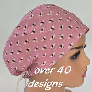 May include: A pink scrub cap with a repeating pattern of yellow and black bees. The cap has white ties and the text "over 40 designs" in white. The cap is on a mannequin head.
