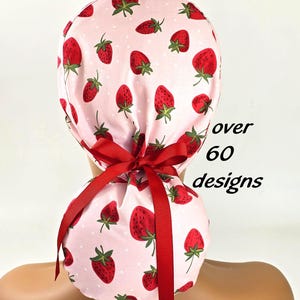 May include: A light pink scrub cap featuring a repeating pattern of red strawberries with green leaves and small white dots. The cap includes a dark red ribbon tied in a bow at the back to secure the fit. The text over 60 designs is visible.