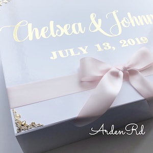 May include: White gift box with a pink ribbon and gold lettering. The box says "Chelsea & John" and "July 13, 2019". The bottom of the box says "Arden Rd".