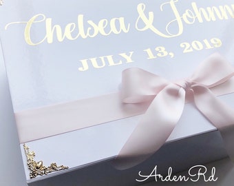 May include: White gift box with a pink ribbon and gold lettering. The box says "Chelsea & John" and "July 13, 2019". The bottom of the box says "Arden Rd".