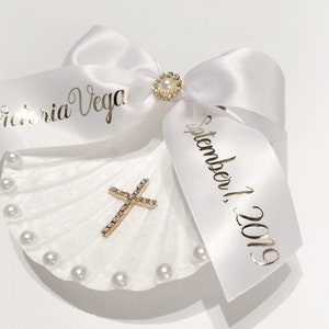 May include: White seashell with a gold cross and pearl accents. A white satin ribbon with gold lettering reads "Victoria Vega September 1, 2019".