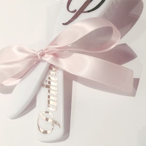 May include: White baby hair brush and comb set with a pink satin ribbon tied in a bow. The comb has the name "Olivia" engraved in gold lettering.