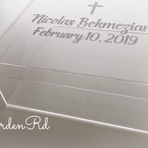 May include: Clear acrylic box with a lid. The lid features a cross and the inscription "Nicolas Bekmezian February 10, 2019" in a script font. The words "Arden Rd" are also visible.