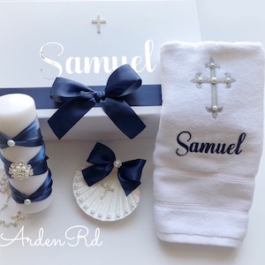 May include: A white gift box with a blue ribbon and the name "Samuel" on the lid. The box is next to a white towel with a silver cross and the name "Samuel" embroidered on it. There is also a white candle with blue ribbon and pearl accents, a white seashell with a blue bow and a silver cross, and a silver rosary.
