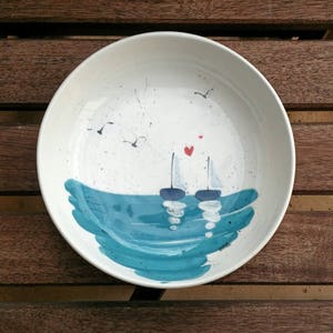 May include: A white ceramic bowl with a blue and white seascape design. Two sailboats are depicted in the ocean with red hearts above them. The bowl has a speckled white glaze.