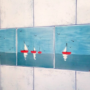 May include: A set of four blue and white ceramic tiles with a nautical theme. The tiles feature a red lighthouse, sailboats, and seagulls against a blue sky and water background.