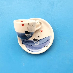 May include: A white ceramic coffee cup with a blue and black abstract design and red hearts. The cup is sitting on a matching saucer.
