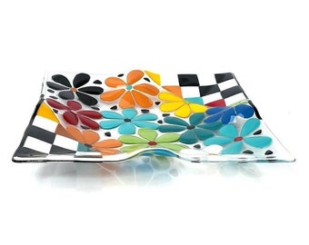 May include: A square glass serving dish with a wavy edge. The dish is decorated with a black and white checkered pattern and colorful flowers. The flowers are in shades of orange, yellow, red, blue, green, and turquoise.