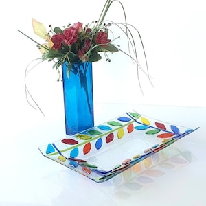 May include: A clear glass serving tray with a colorful leaf design. The tray is decorated with red, green, blue, yellow, and orange leaves. A blue glass vase with a bouquet of red roses is in the background.