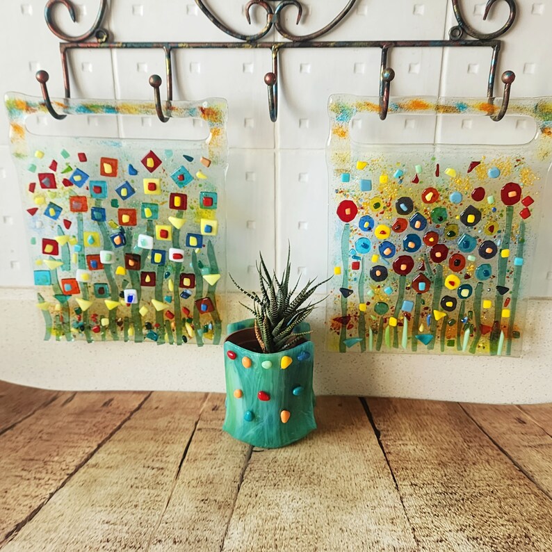 Op de afbeelding: Twee kleurrijke glazen panelen met abstracte bloemmotieven hangen aan een muur met haken. Een kleine groene pot met een vetplant staat voor de panelen.