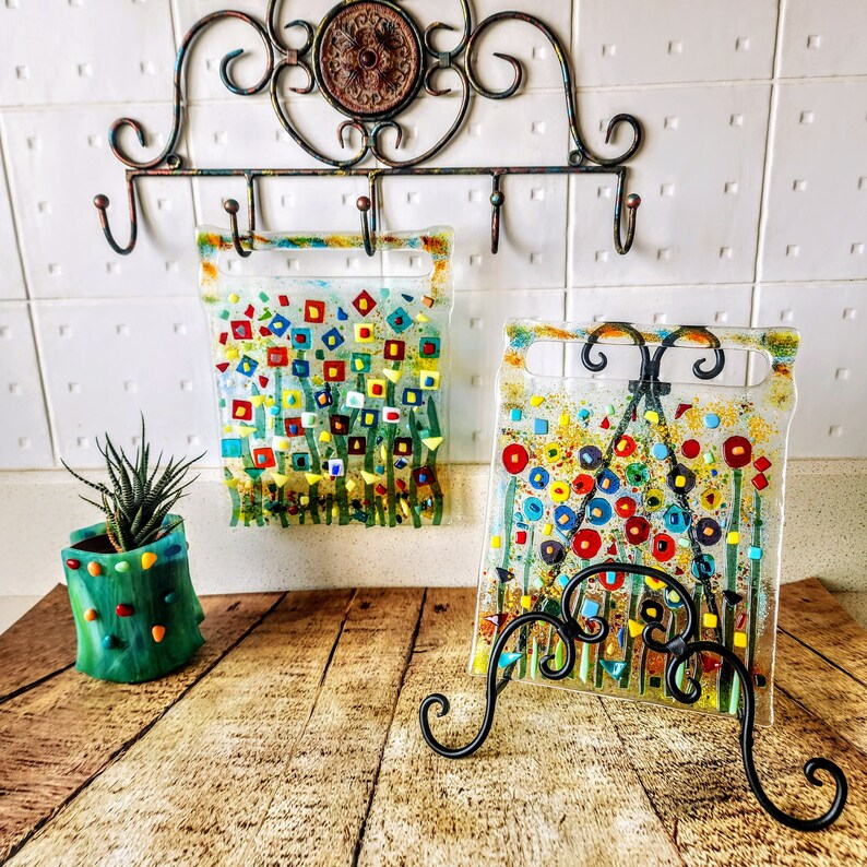 Op de afbeelding: Twee kleurrijke glazen kunstwerken met abstracte bloemmotieven, een op een zwarte metalen ezel. De kunstwerken bevatten een mix van rode, gele, blauwe en groene kleuren.